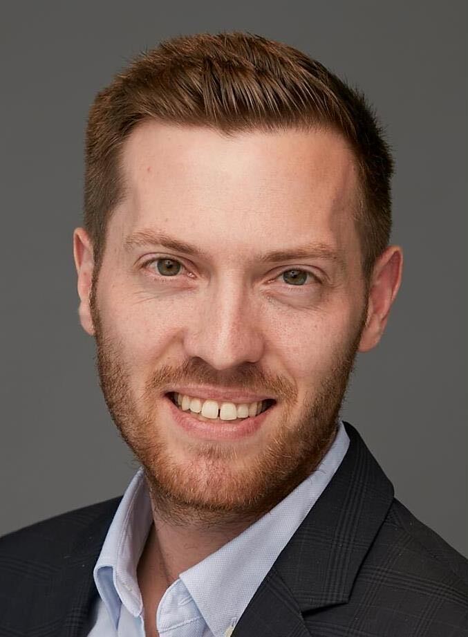 Smiling man with short brown hair and beard wearing a light blue shirt and dark blazer against a gray background.
