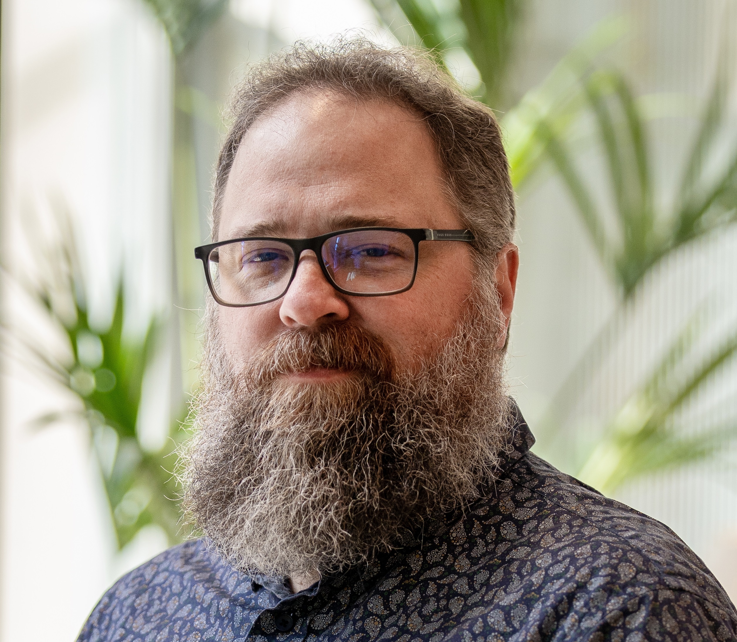 Bearded man with glasses wearing a patterned shirt, standing indoors with green plants in the background.