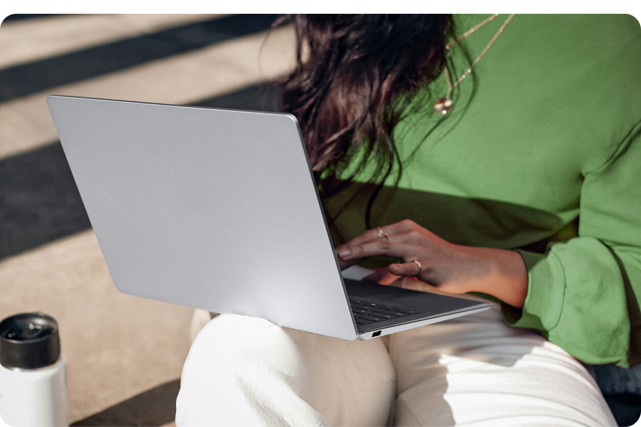 Person wearing green top and white pants typing on a slim silver laptop outdoors with a water bottle nearby.