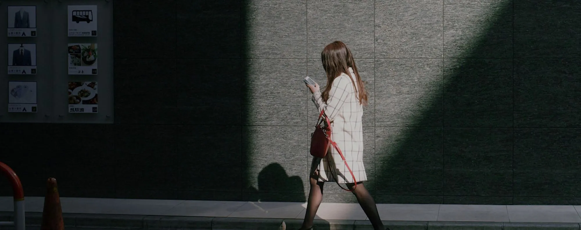 Woman in a white checkered coat walking past a dark tiled wall while looking at her phone.