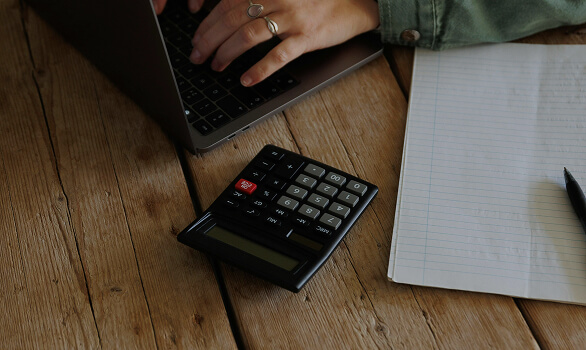 Person typing on a laptop keyboard at a wooden table with a calculator and a lined notebook with a pen.
