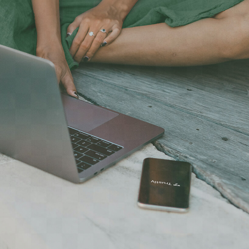Person sitting cross-legged on a wooden surface using a laptop with a smartphone nearby displaying the Trustly logo.