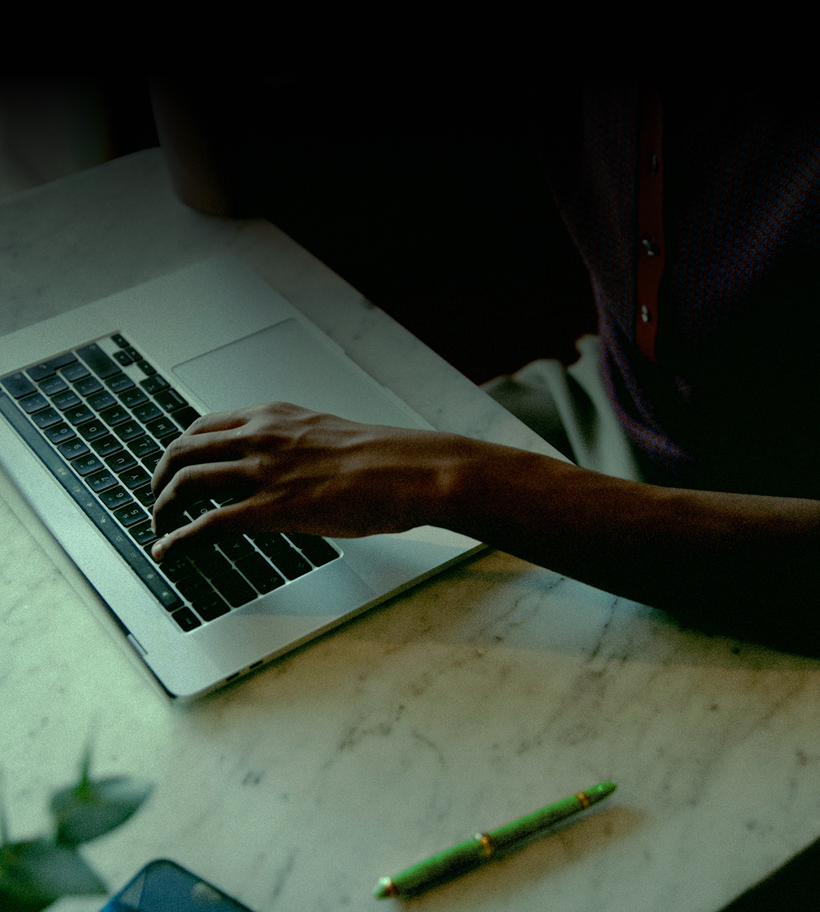 Person typing on a laptop keyboard on a marble table with a green pen nearby.