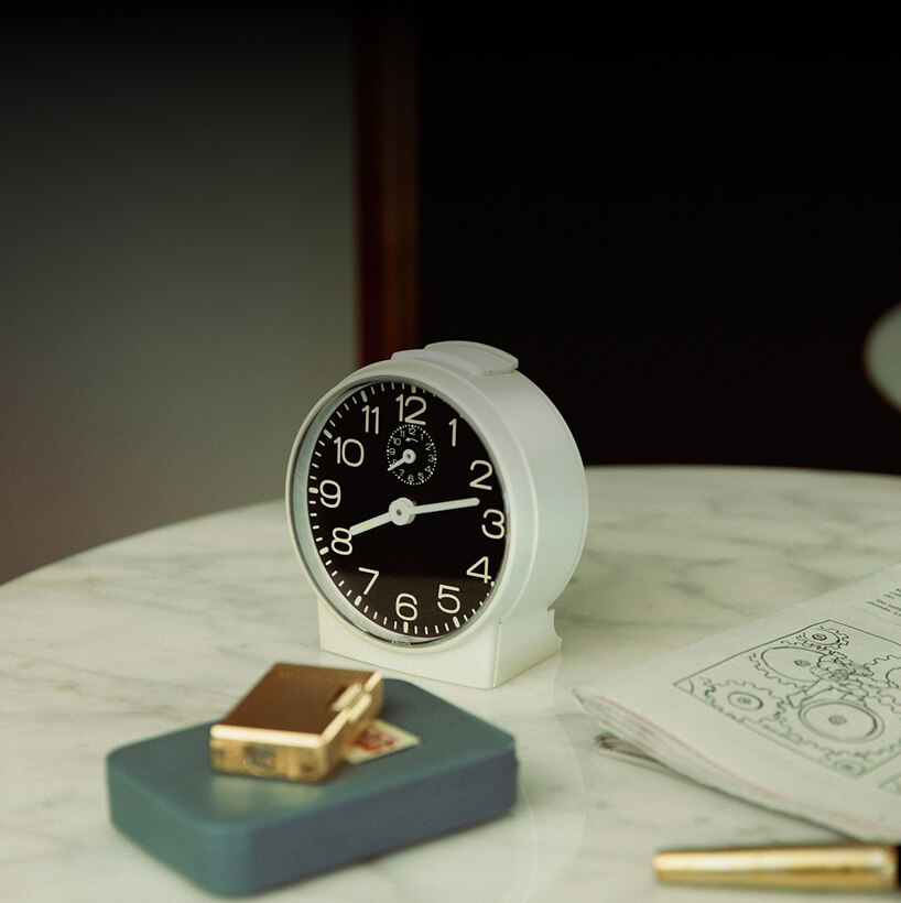 White analog alarm clock on marble table alongside a blue case with a gold lighter and an opened book with mechanical drawings.