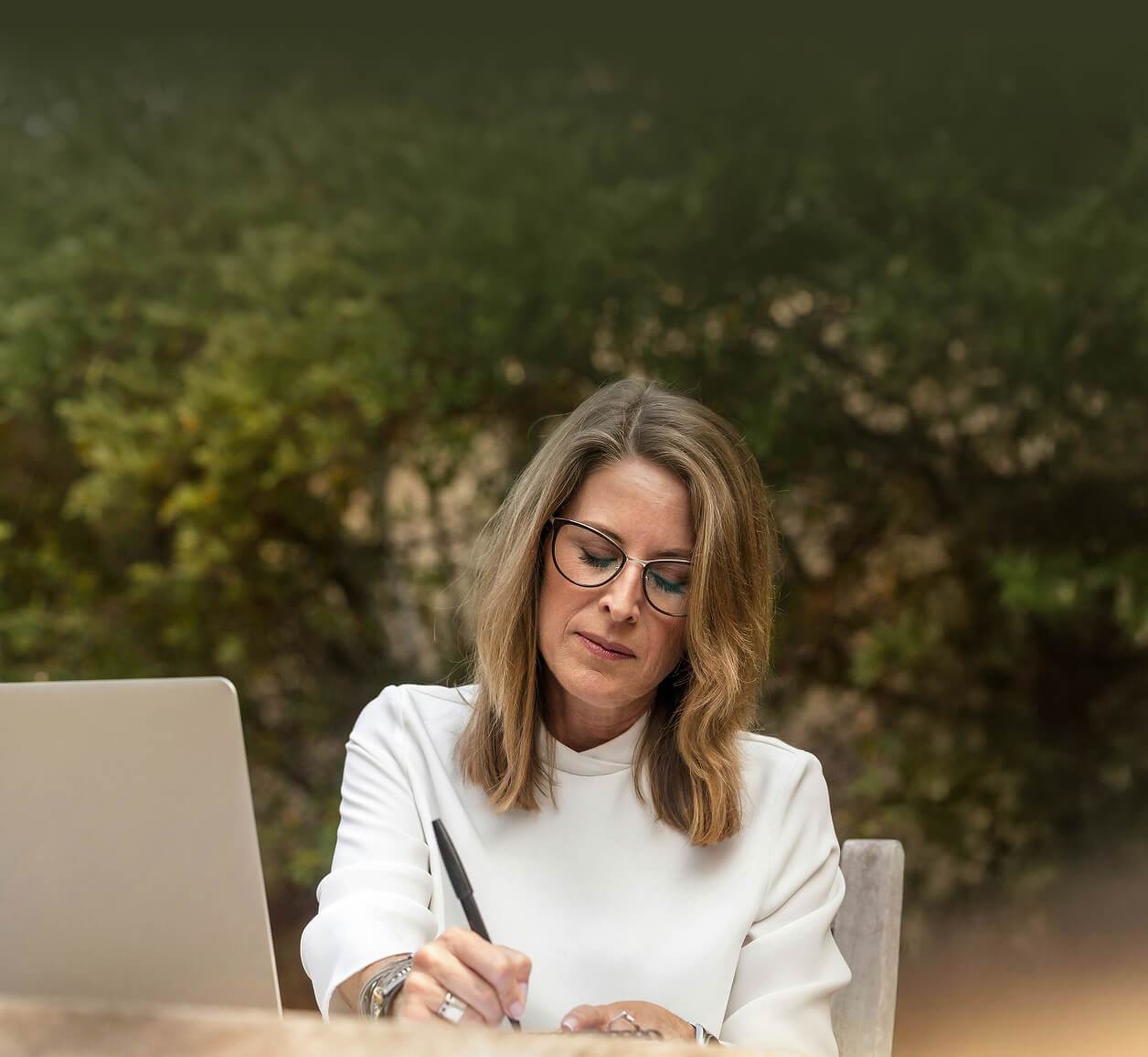 Woman with glasses and shoulder-length blonde hair writing with a pen outdoors next to a laptop.