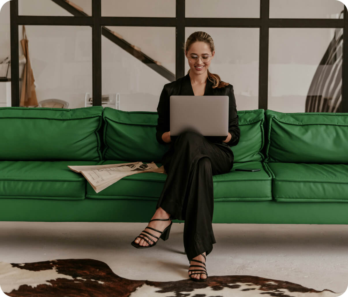 Woman in black outfit sitting on a green couch using a laptop, with a newspaper and smartphone beside her.
