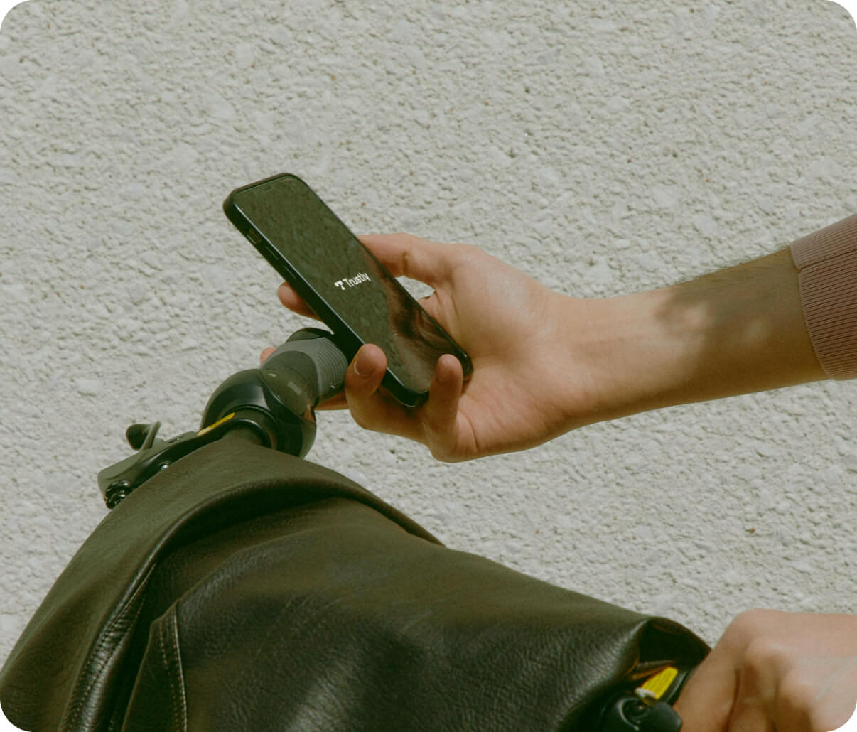 Hand holding a smartphone with the Trustly logo, near the handlebars of a black leather bag on a textured wall background.
