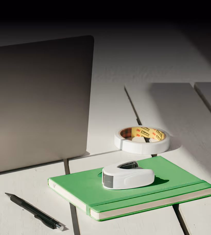 White desk with a closed laptop, green notebook with a white stapler on top, roll of white tape, and a black pen.