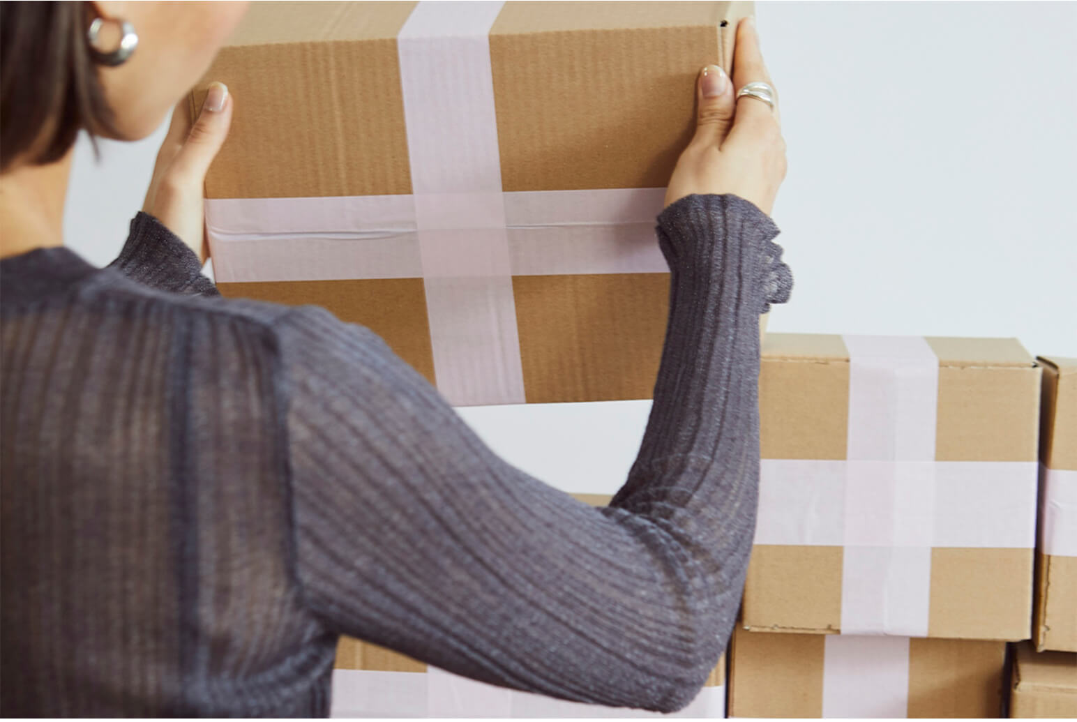 Person holding a cardboard box sealed with white tape in front of a stack of similar boxes.