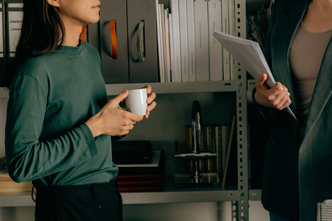 Two people having a discussion near shelves filled with binders and office supplies, one holding a white coffee mug and the other holding a stack of papers.