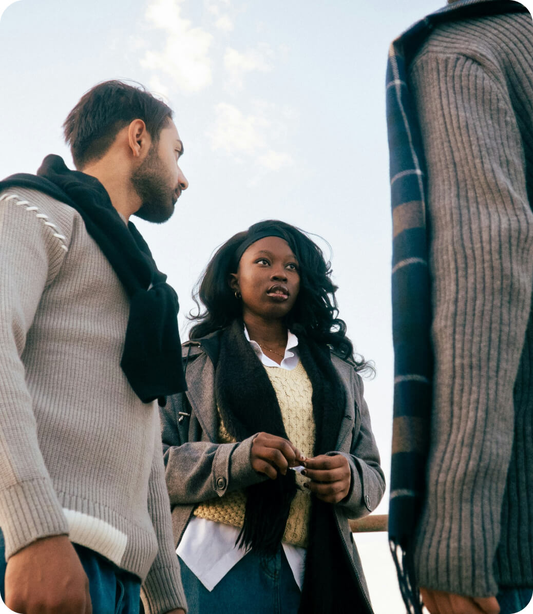 Three people standing outdoors having a conversation, dressed in sweaters and scarves under a partly cloudy sky.