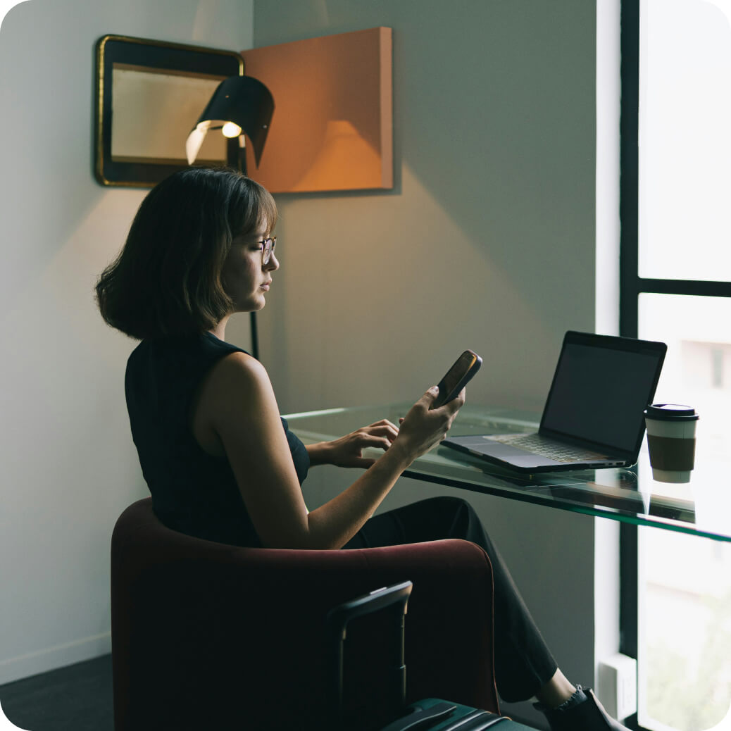 Woman sitting in a chair using a smartphone with a laptop and coffee cup on a glass desk by a window.