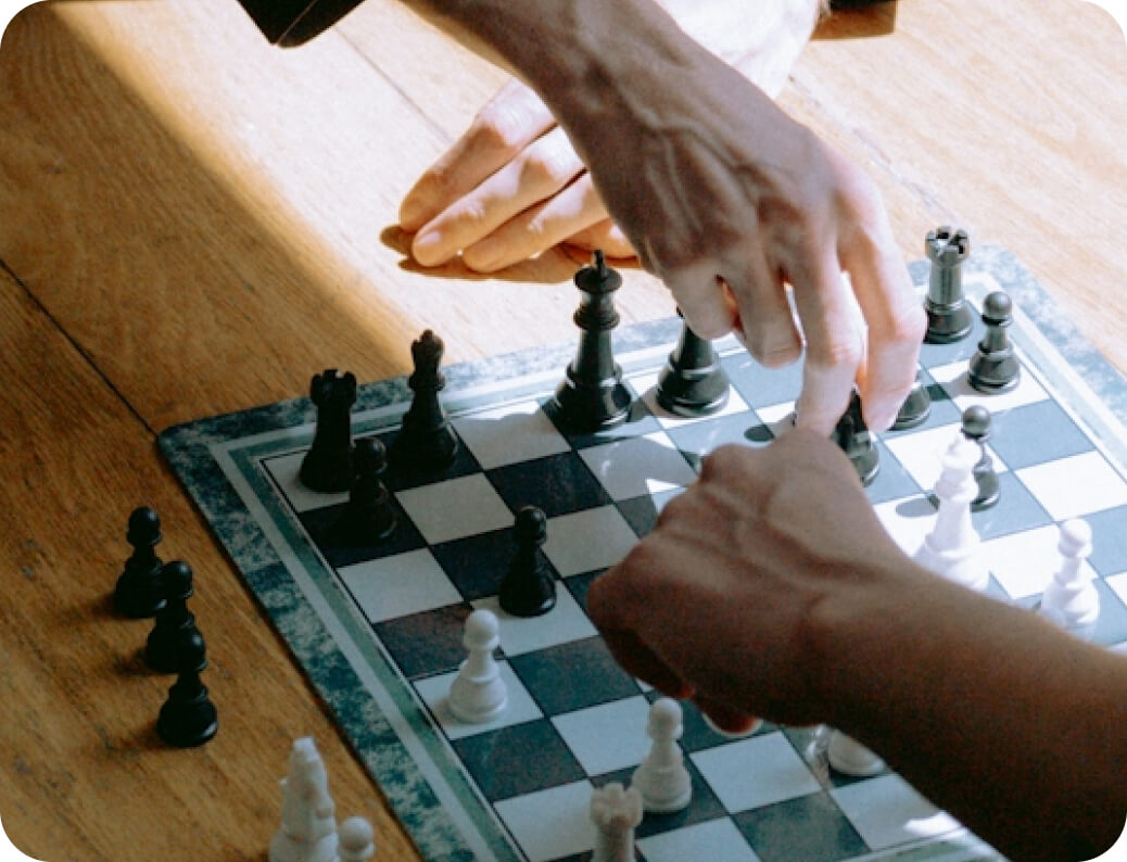Two people playing chess on a wooden table, with one hand moving a black chess piece.