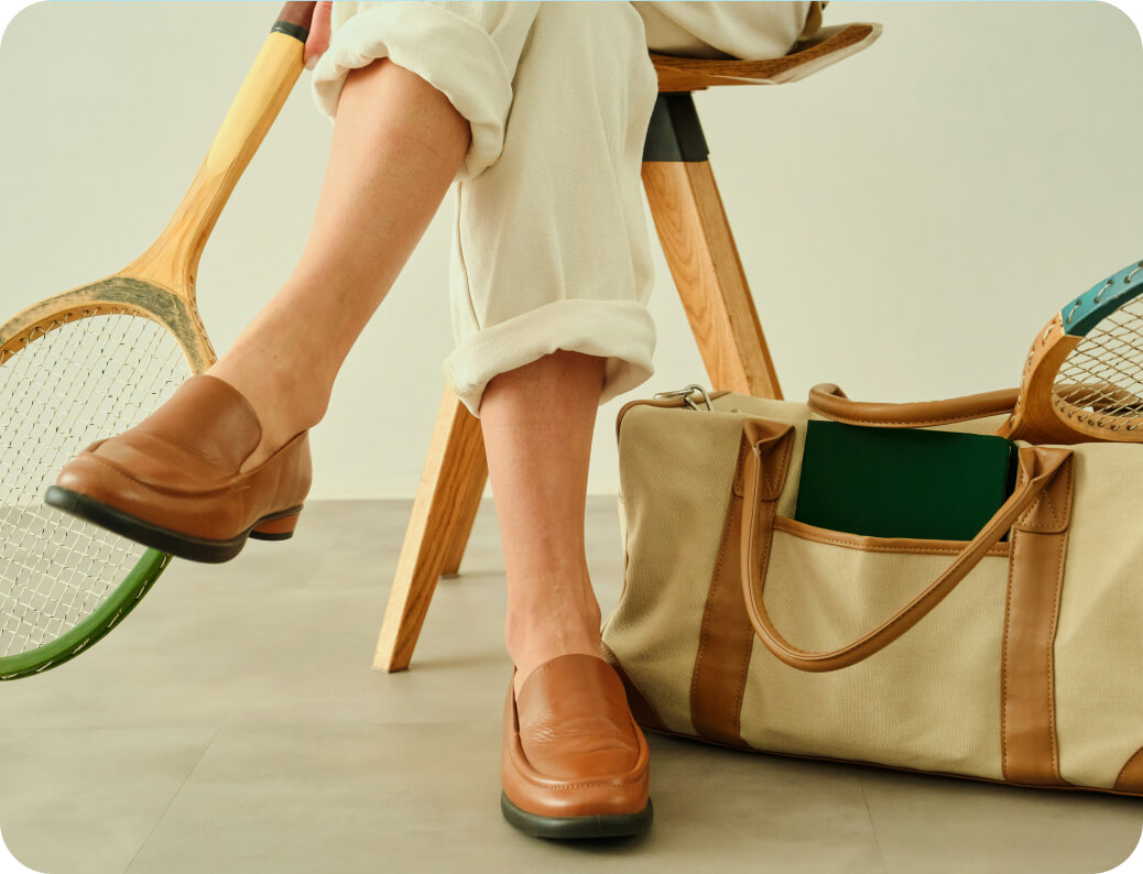 Person sitting on wooden chair with rolled-up cream pants and brown loafers, holding a vintage wooden tennis racket next to a beige canvas and leather sports bag with another tennis racket inside.