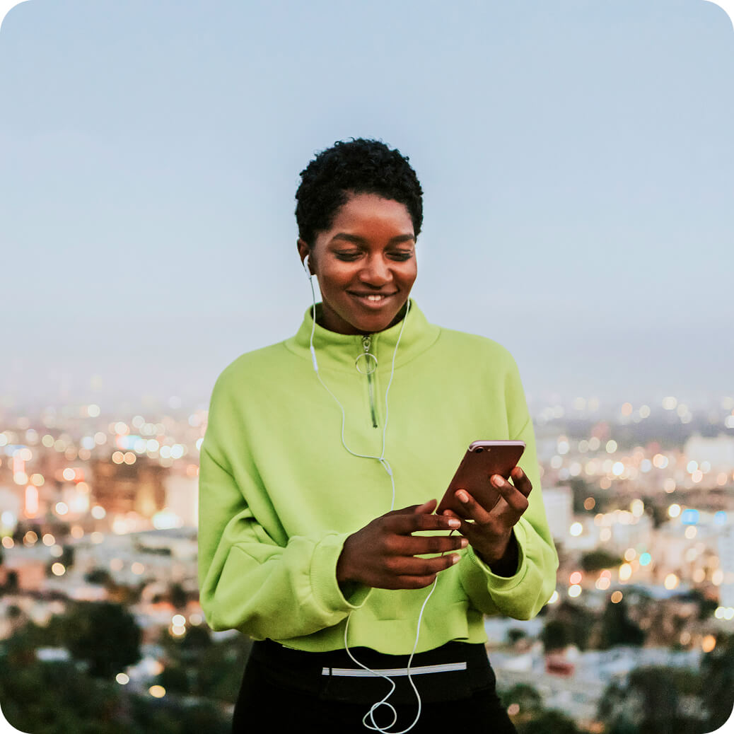 Smiling young woman in a green sweatshirt listens with earphones while holding a smartphone against a cityscape background at dusk.