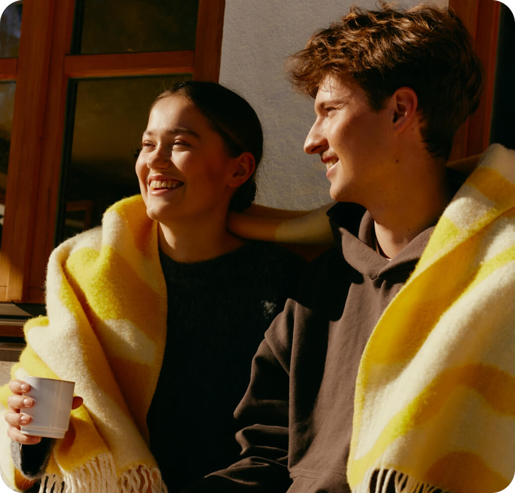 Young woman and man sitting close together, wrapped in a yellow and white blanket, smiling in warm sunlight.