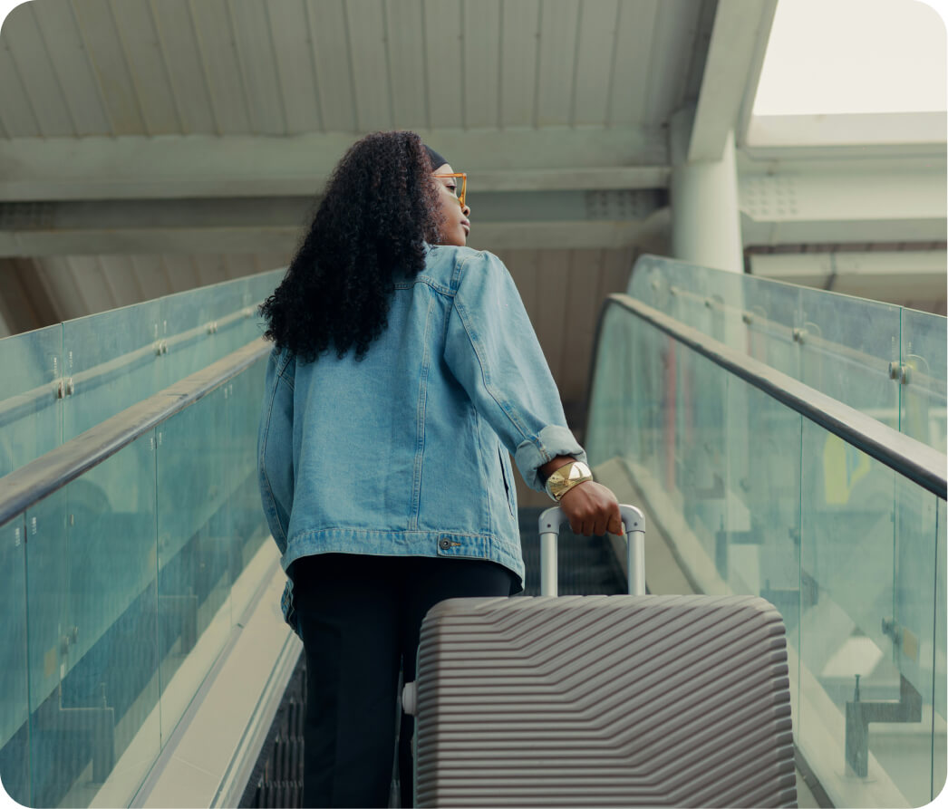 Woman with curly hair wearing a denim jacket and sunglasses, holding a large suitcase on an escalator.