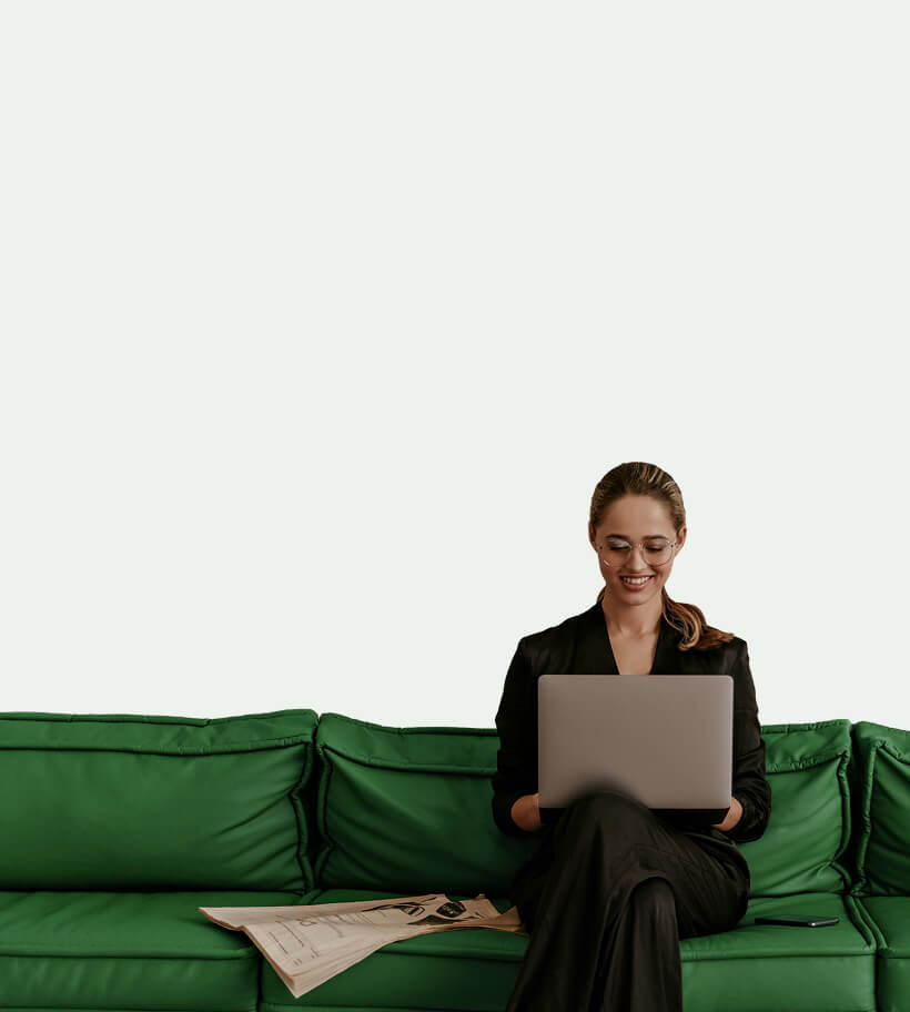 Smiling woman in glasses and black outfit sitting on a green couch, using a laptop with a newspaper and smartphone beside her.