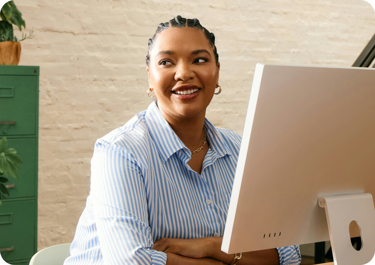 Smiling woman with braided hair and gold hoop earrings sitting at a desk in front of a computer.