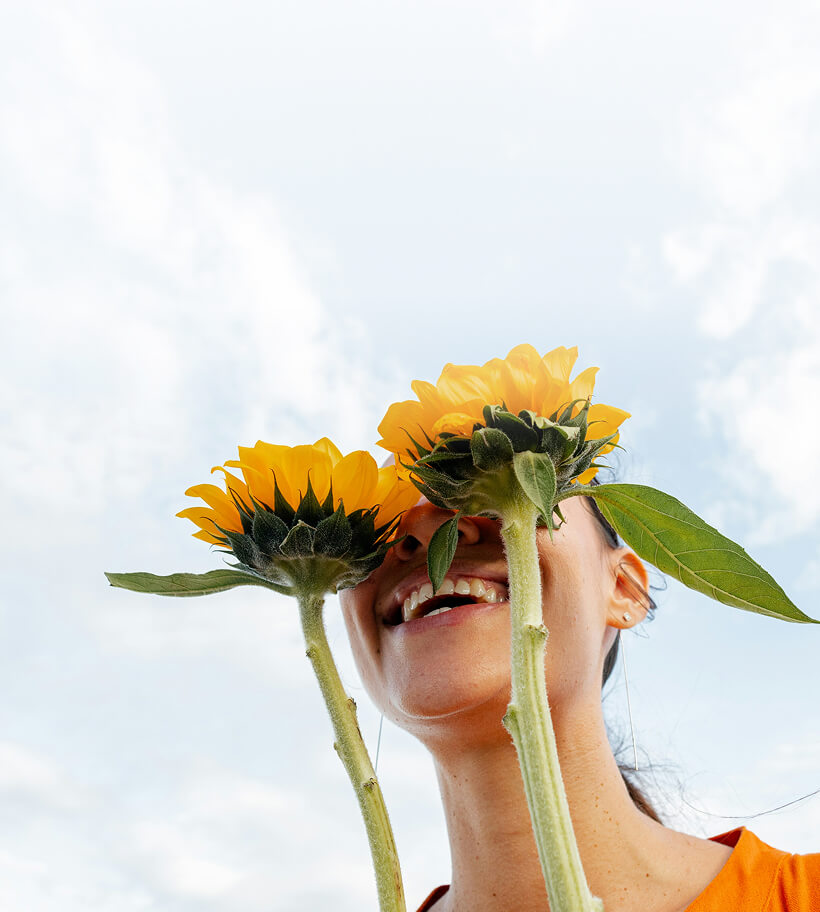 Woman smiling holding two sunflowers in front of her eyes against a cloudy sky.