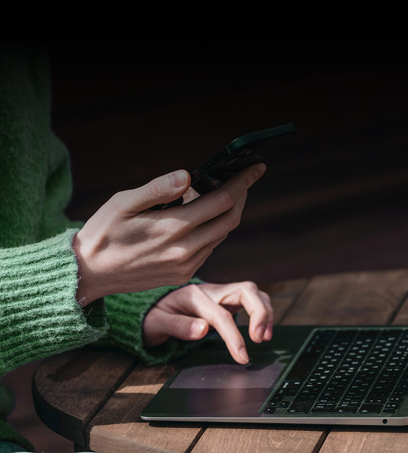 Person wearing a green sweater using a smartphone in one hand and typing on a laptop with the other hand on a wooden table.