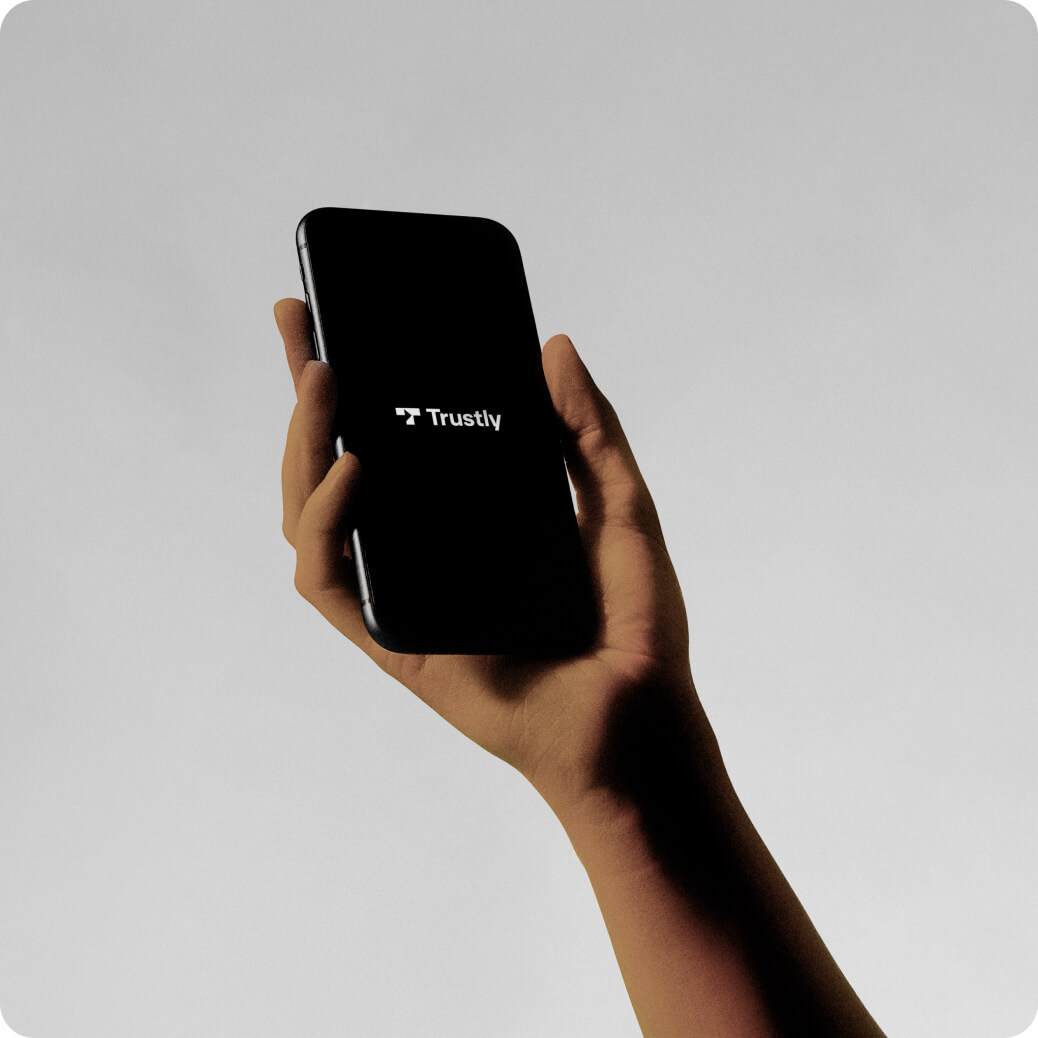 Hand holding a smartphone displaying the Trustly logo on a black screen.