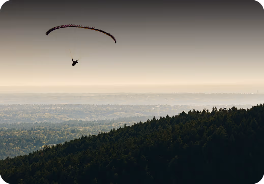 Person paragliding above a densely forested hillside under a hazy sky.