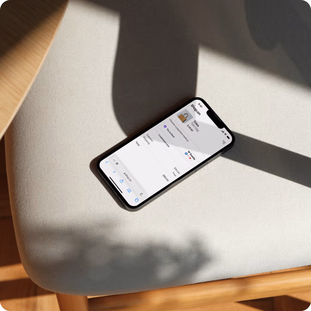 Smartphone on a light fabric chair seat displaying a checkout screen for a $24.99 tote bag with payment options.