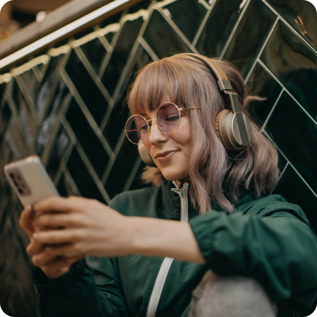 Woman with light brown hair wearing round glasses and headphones, smiling while holding a smartphone.