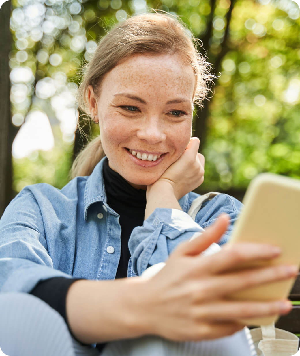 Smiling young woman with freckles sitting outdoors and looking at her smartphone.
