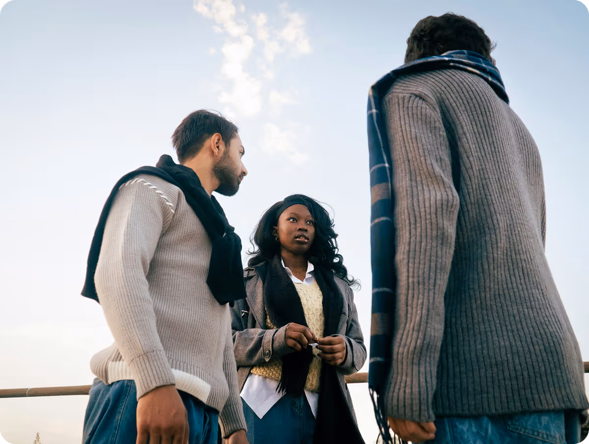 Three young adults in sweaters and scarves standing outdoors under a clear sky, engaged in conversation.