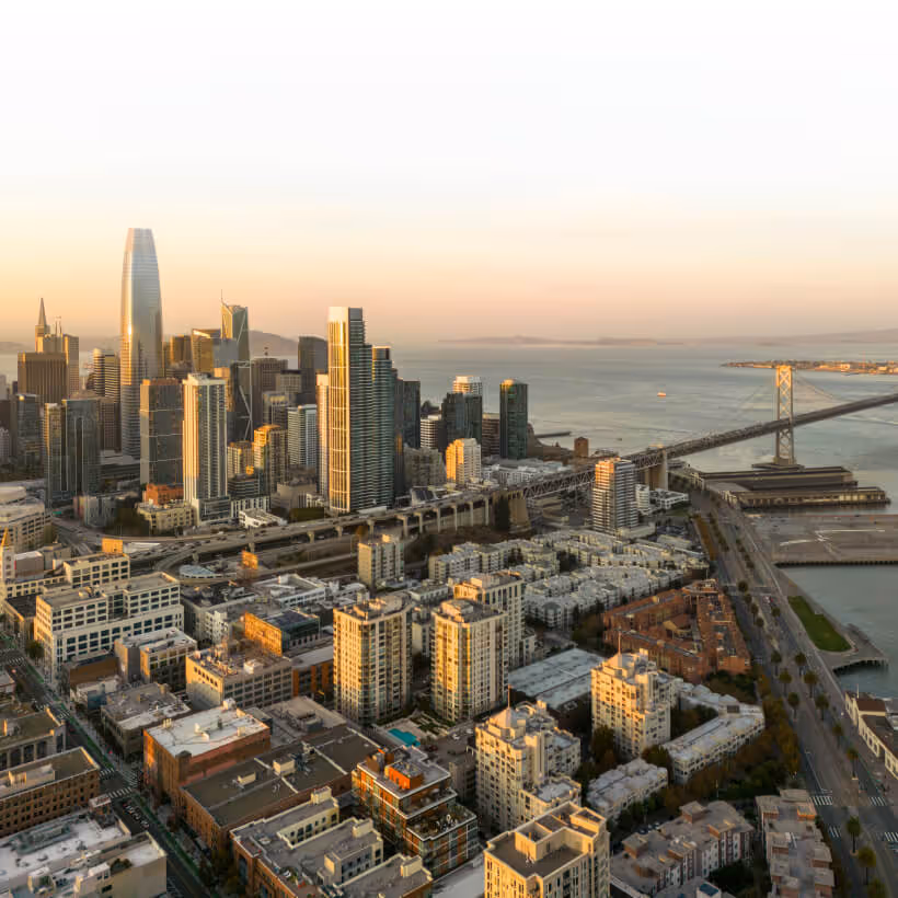 Aerial view of downtown San Francisco with skyscrapers, residential buildings, and the Bay Bridge over the water at sunset.