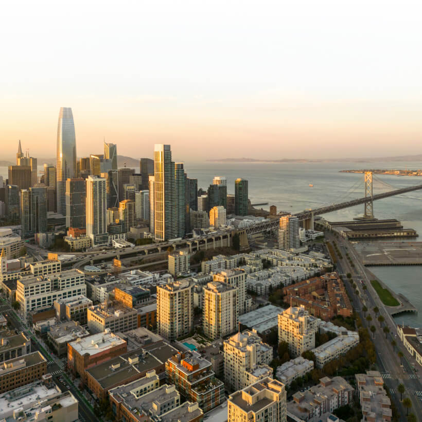 Aerial view of downtown San Francisco with skyscrapers, residential buildings, and the Bay Bridge over the water at sunset.
