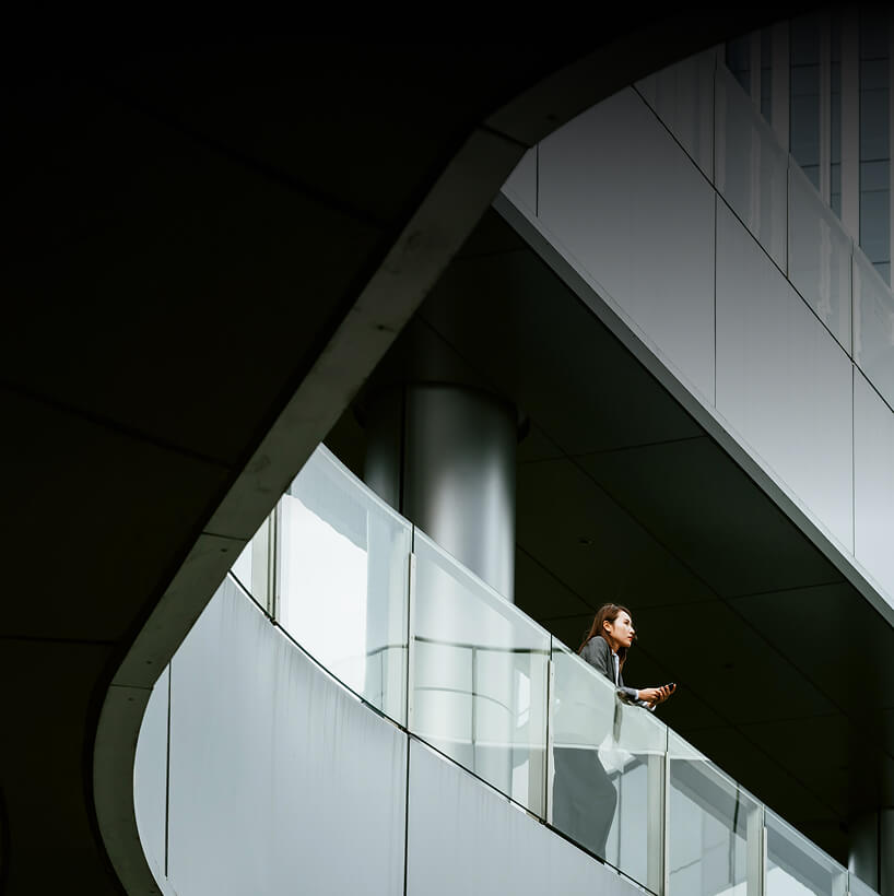 Woman standing on a modern building balcony holding a smartphone, looking contemplatively.