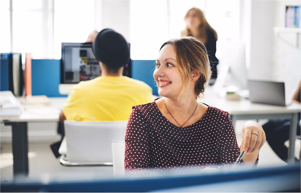 Smiling woman in polka dot blouse sitting at desk in office, with coworkers and computer screens in background.