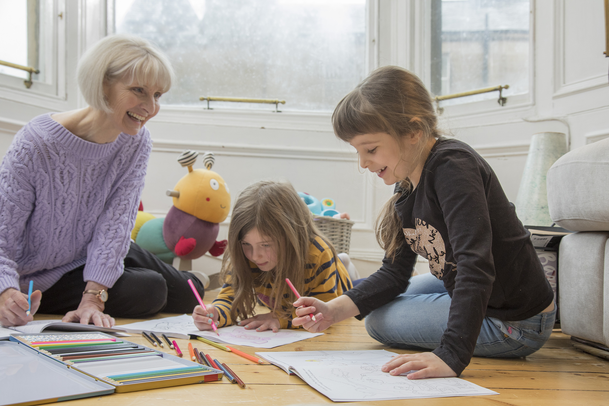 Smiling elderly woman and two young girls color together on a wooden floor near a window with colored pencils and coloring books.