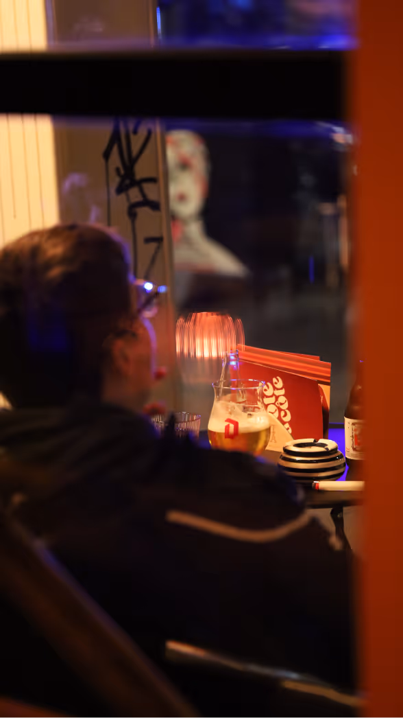 Persoon kijkt via raam naar binnen in een café met een glas bier en een gestreept bord op tafel.