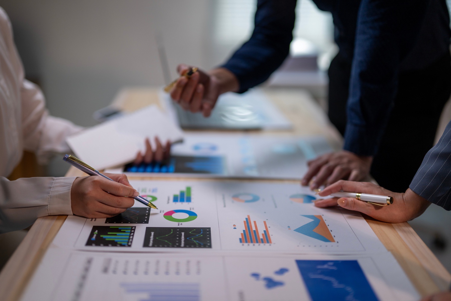 Close-up of three people analyzing financial charts and graphs on a desk, pointing with pens.