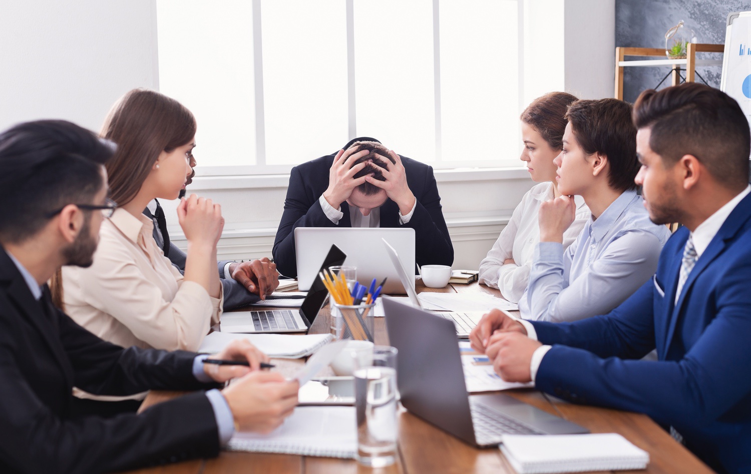 A stressed businessman with head in hands sits at a conference table, surrounded by colleagues who watch him attentively.