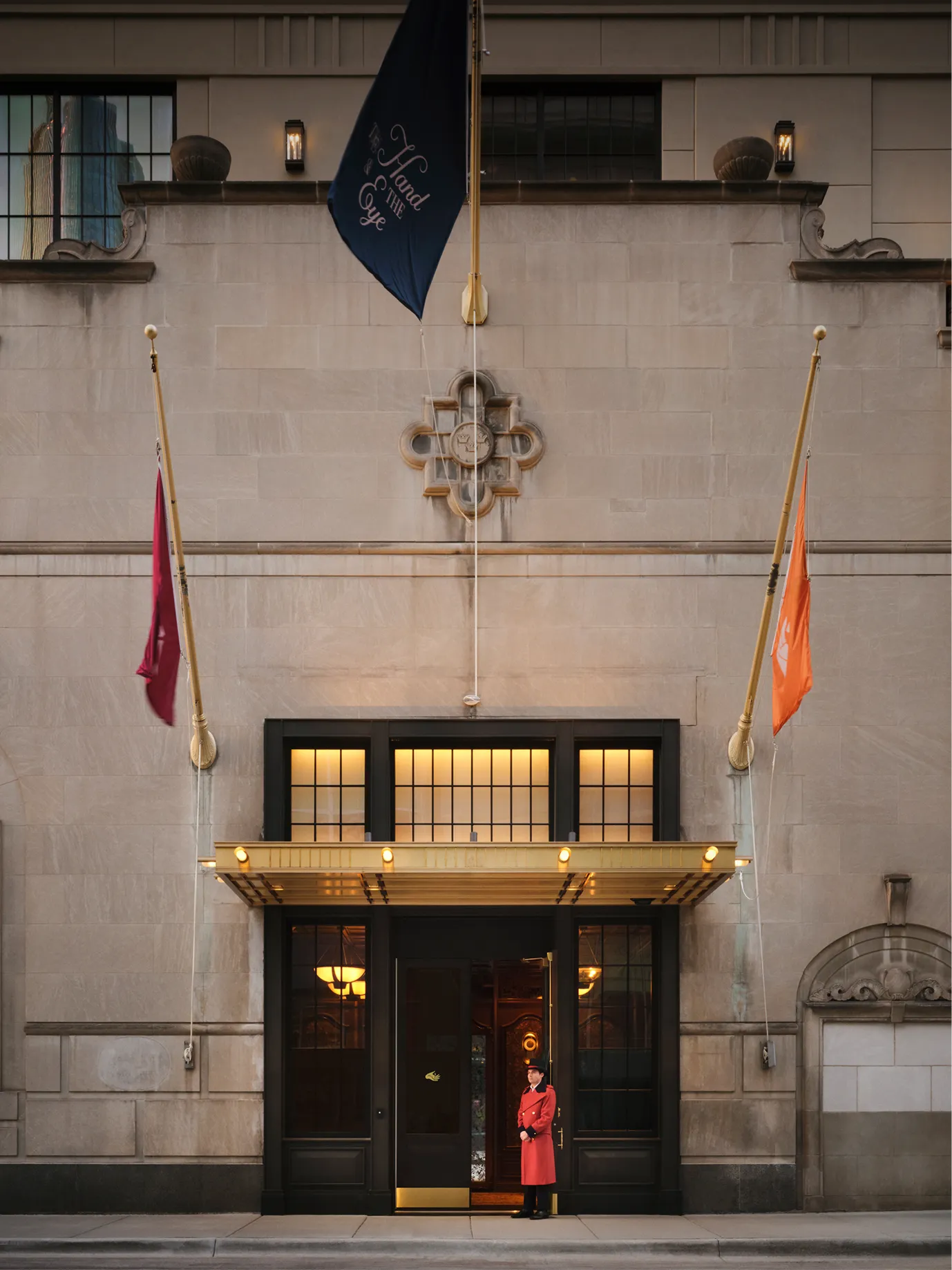 Doorman in red coat standing outside an elegant stone building entrance with flags and lit canopy.