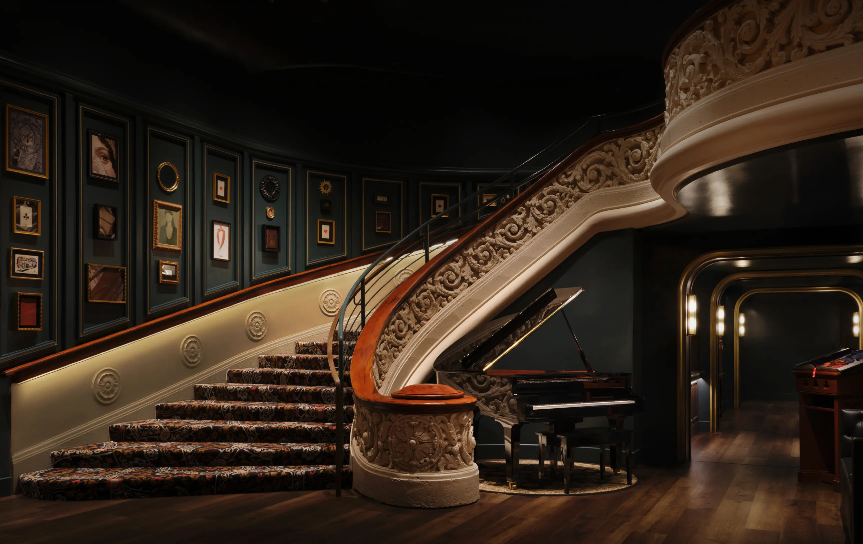 Ornate curved staircase with patterned carpet and framed art on walls beside a black grand piano.