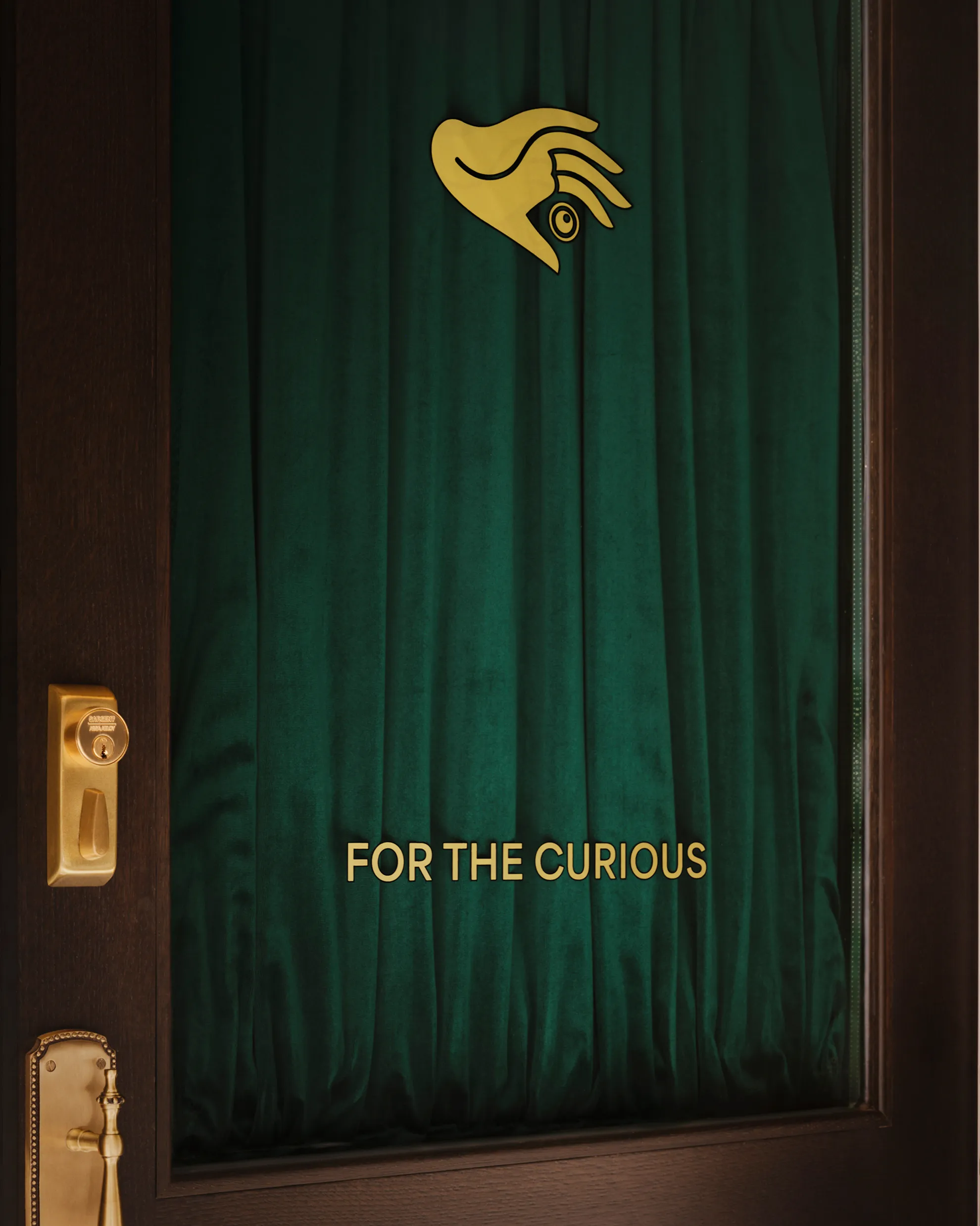 Dark wooden door with gold lock, a green curtain, gold hand symbol, and text 'FOR THE CURIOUS'.