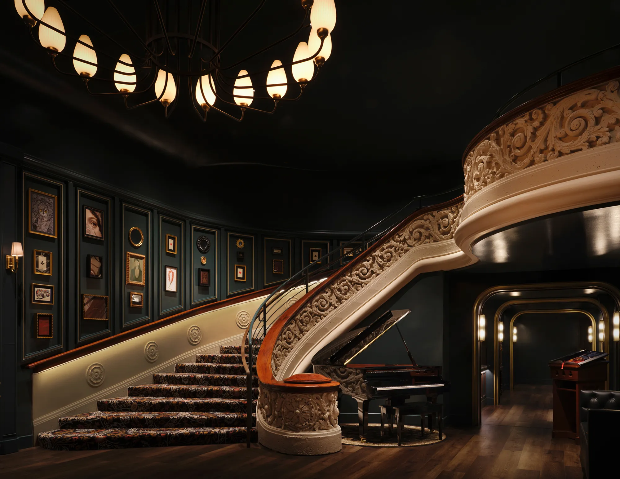 Grand curved staircase with ornate railings and framed art on walls, a grand piano beneath, and warm lighting.