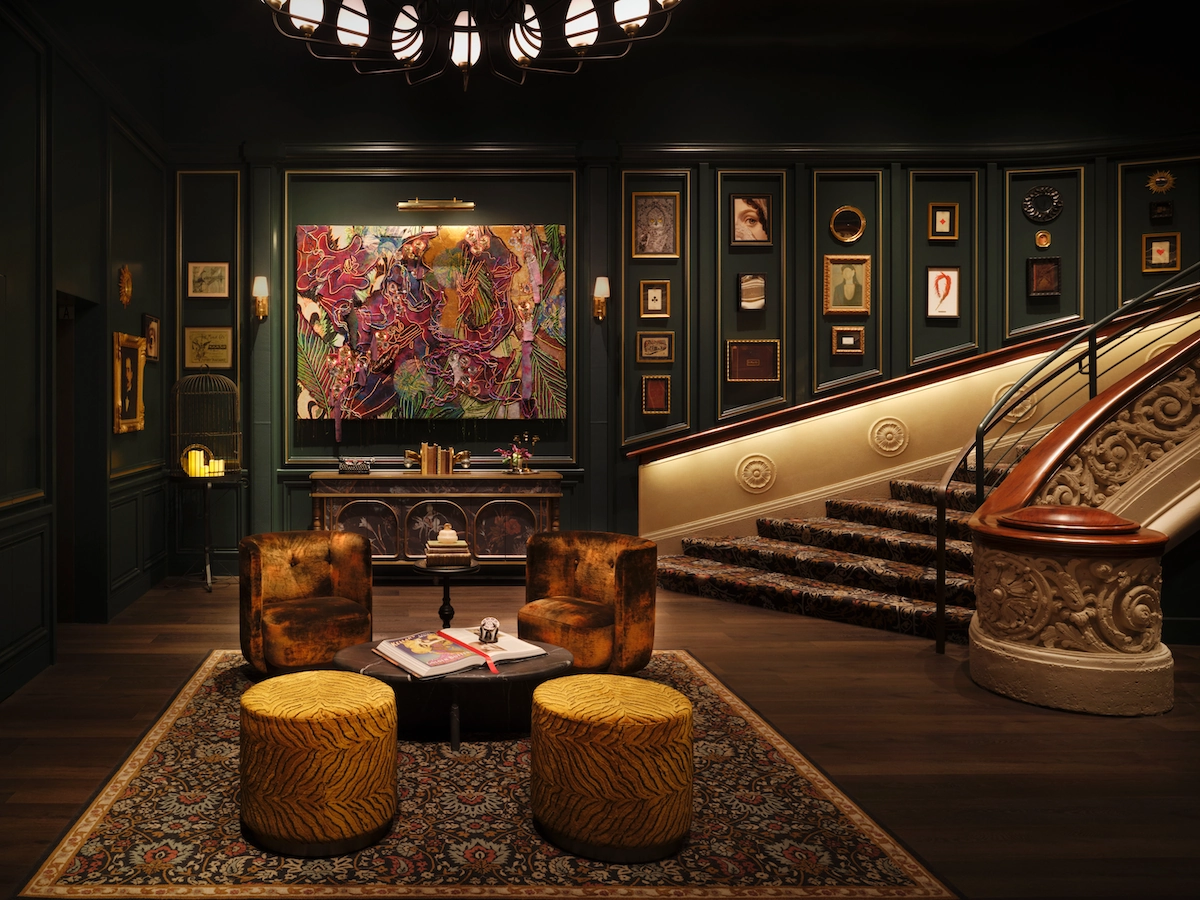 Luxurious lounge with patterned rug, two brown chairs, yellow stools, and staircase with framed art on walls.