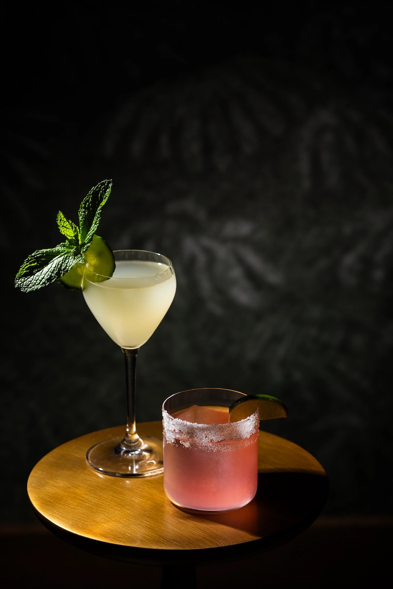 Two cocktails on a wooden round table: a pale drink garnished with mint, and a pink drink with salted rim and lime wedge.