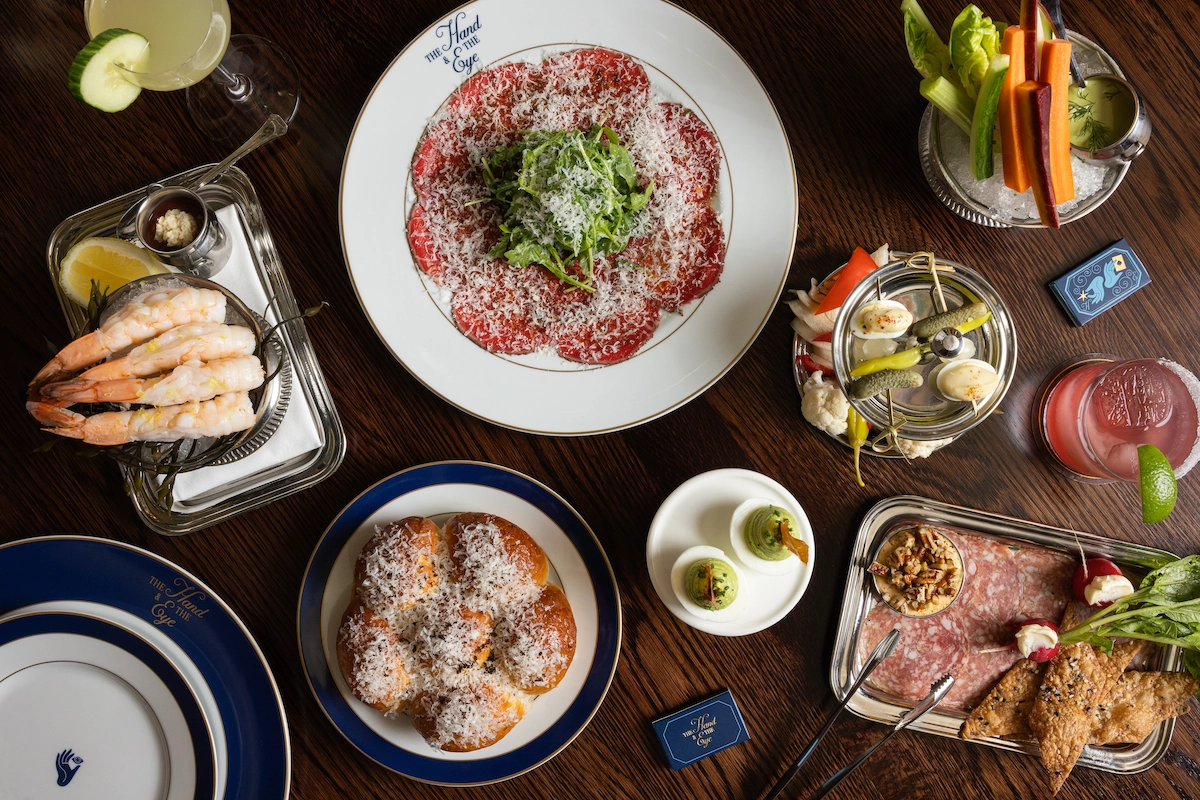 Overhead view of assorted starters including shrimp, beef carpaccio, deviled eggs, veggies, and breads on a wooden table.