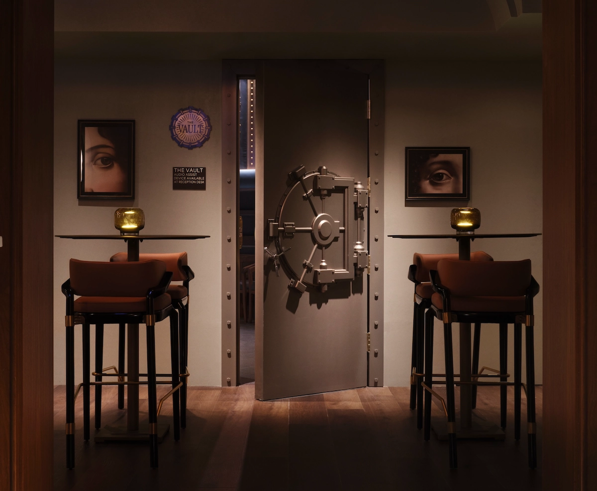Dimly lit room with a heavy vault door slightly open, two tables with chairs and eye-themed art on walls.