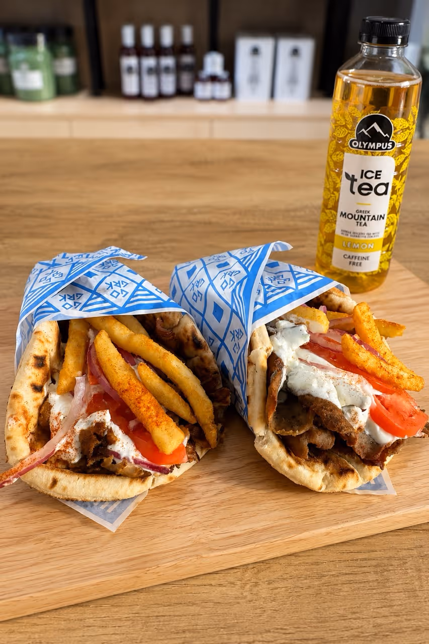 Two Greek gyros wrapped in pita with fries, tomato, onion, and tzatziki, next to a bottle of lemon iced tea.