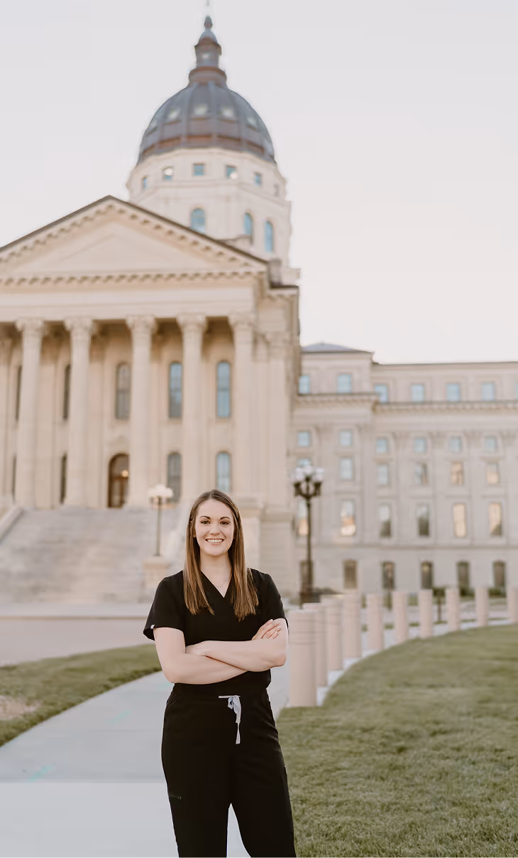 Smiling woman in black scrubs standing with arms crossed in front of a large domed government building.