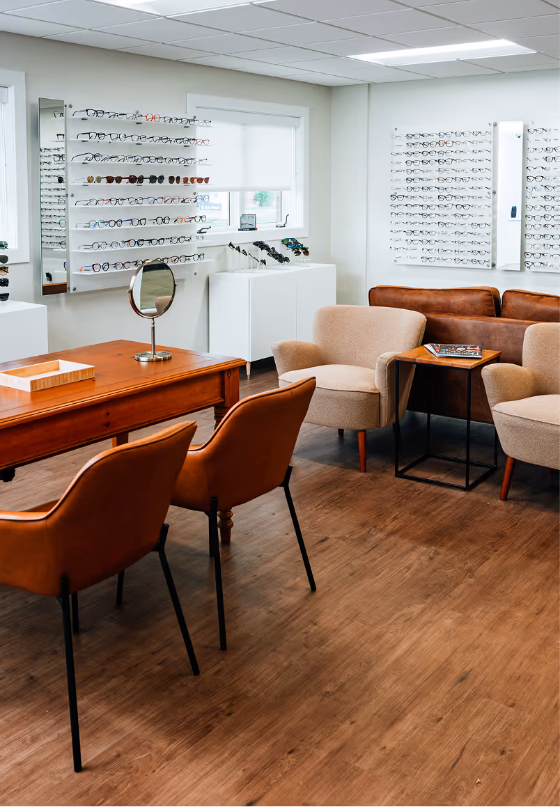 Modern eyewear store interior with display walls of glasses, brown chairs around a wooden table, beige armchairs, and a brown leather sofa.